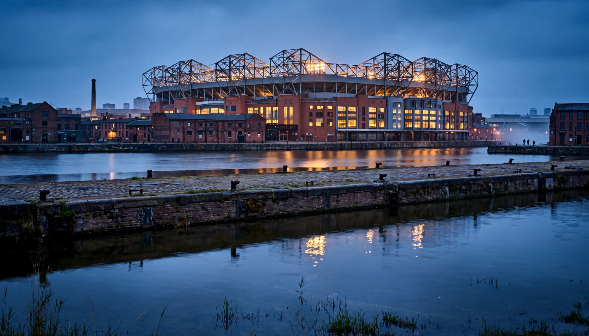 A glowing football stadium stands beyond a canal at dusk with reflections in the water.