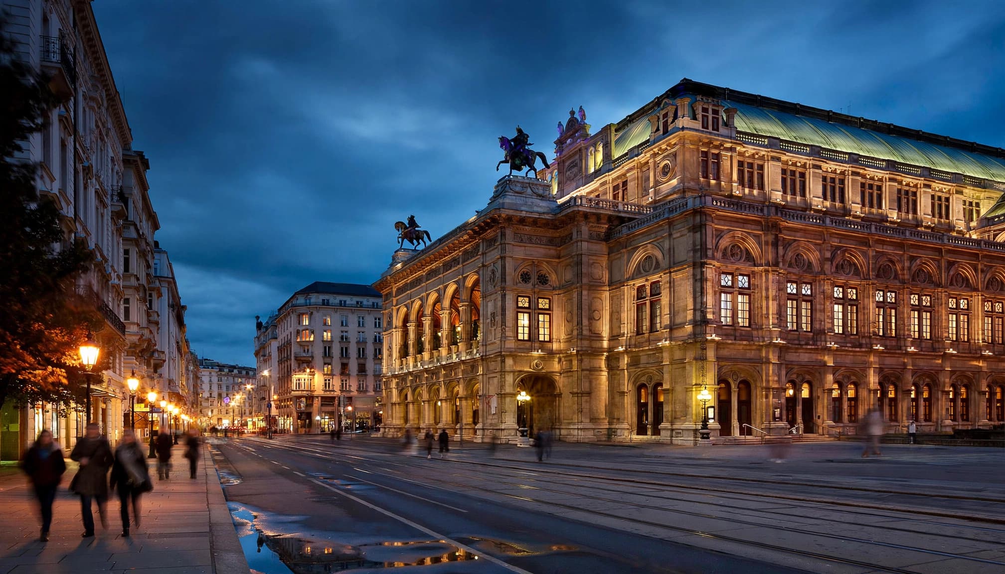 The Vienna State Opera glows across the street as pedestrians pass at dusk.