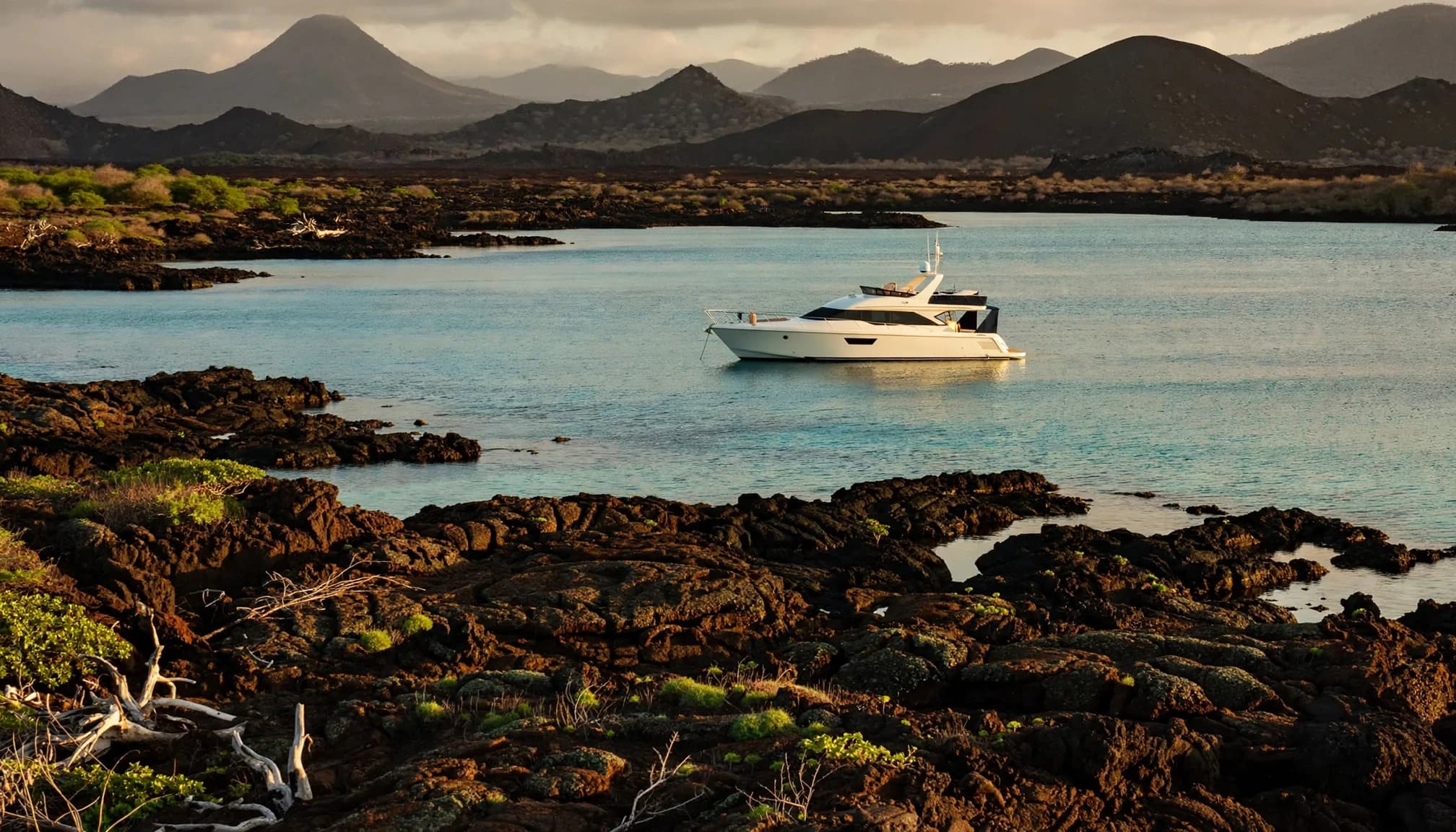 A white yacht anchored in turquoise water near a volcanic shoreline with dark peaks in the background.