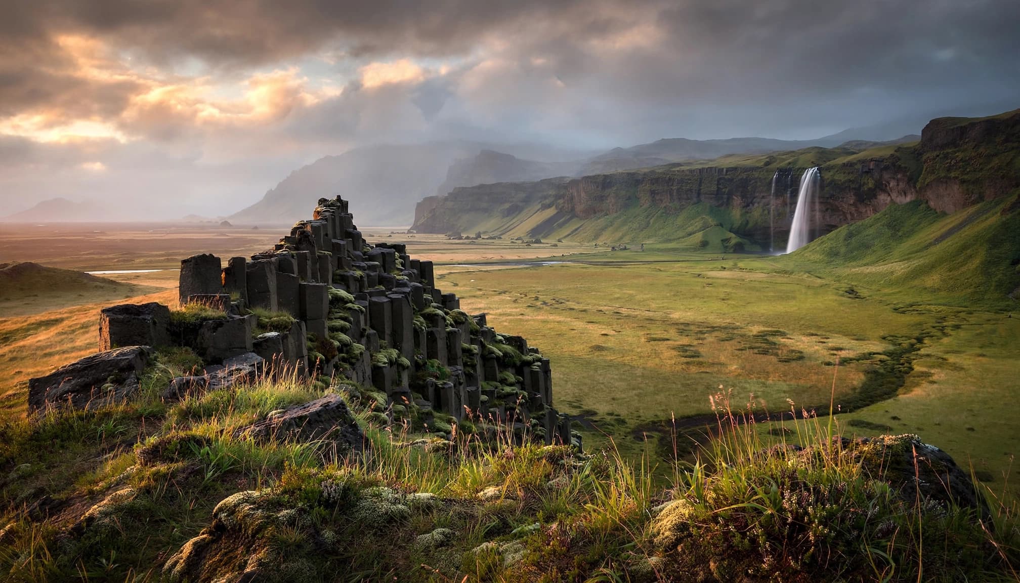 Wide landscape view of an Icelandic valley with basalt columns in the foreground and a waterfall in the distance.