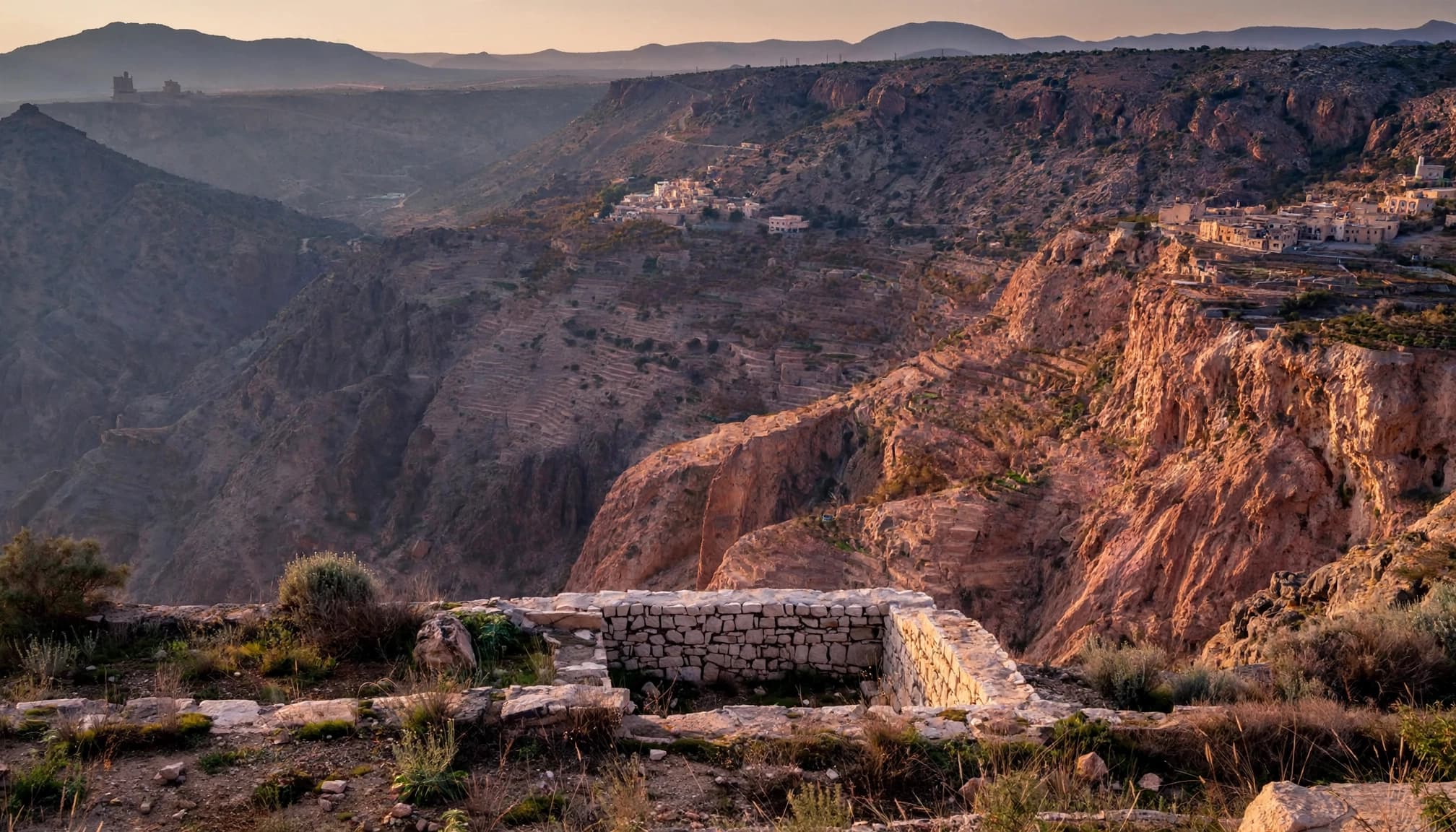 A stone terrace overlooks rose-colored cliffs, terraced villages, and distant mountains in Oman.